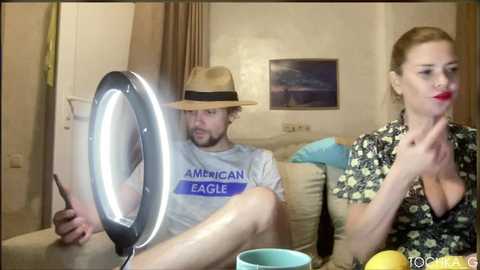 A video captures a man with a hat, a woman with red lipstick, and a ring light in a living room.