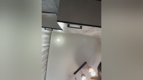 Video of a modern, dimly-lit room with white walls, exposed concrete ceiling, and black metal lighting fixtures. A small, circular ceiling light emits a soft glow. The room has a minimalist design with no visible furniture or decor.