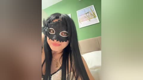 Video of a woman in a black lace masquerade mask with large eye holes, wearing a black lace dress, standing in a room with a green wall and a beige headboard.
