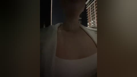 A dimly lit video of a woman with fair skin, wearing a white tank top, partially obscured by shadows. Background shows a window with blinds and a brick wall.