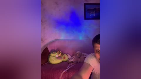 Video of a dimly lit bedroom with a young boy, wearing glasses, lying on a bed with a stuffed toy and a blanket, under a blue glow.