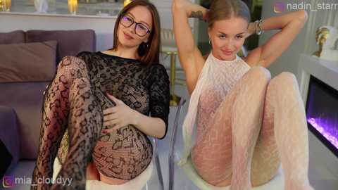 Video of two young women in a modern living room: one in a sheer black lace dress, the other in a sheer white dress, both seated with legs crossed.