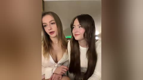 Video of two young women with long hair, one with light brown hair and another with dark brown hair, wearing white tops, looking at a mirror, creating a playful and intimate moment.