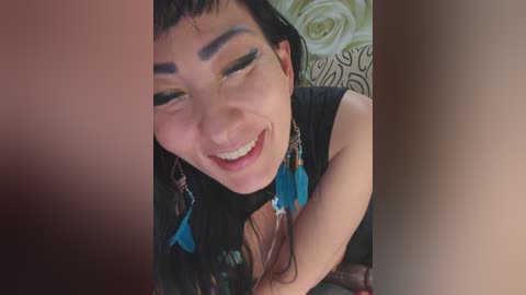 Video of an Asian woman with long black hair, wearing blue feather earrings, smiling with closed eyes, lying on a bed with green floral sheets.