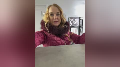 Video of a blonde woman with wavy hair in a pink quilted jacket, leaning over a table in a blurred, dimly lit room with a sign saying \"Coffee\" in the background.