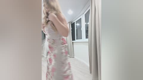 A blurred video of a woman in a white floral dress with long hair, standing in a modern, minimalist hallway with large windows and light wooden floors.