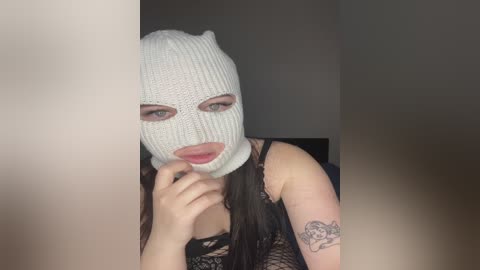 Video of a young woman with light skin, long dark hair, wearing a white knitted balaclava, a black lace top, and a tattoo of a woman on her arm, indoors, dim lighting.