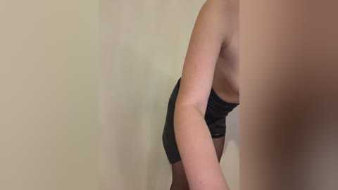 Video of a light-skinned person in a black, strapless top, leaning forward. The background is a plain beige wall. The image focuses on the upper body and torso.