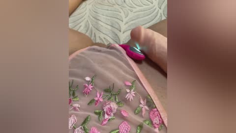 A close-up video of a woman's hand holding a small, bright pink vibrator against her sheer, embroidered pink underwear featuring floral designs. The background shows a textured, light green pillow.