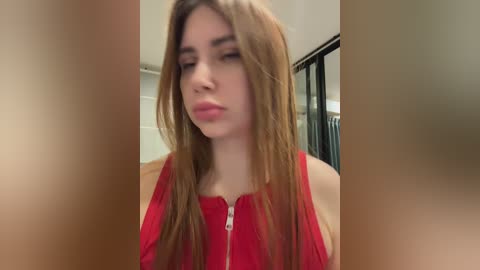 Video of a young woman with long, straight, light brown hair and fair skin, wearing a red, sleeveless top, making a pouty face. Background features a beige wall and a glass door.