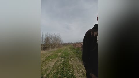 A video of a man standing in a foggy, rural field. He wears a dark jacket, and the background features leafless trees and a cloudy sky. The scene is muted and moody.