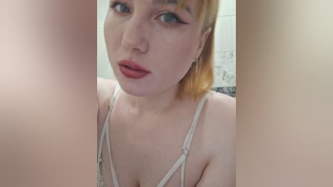 Video of a fair-skinned, blue-eyed woman with short, light brown hair, wearing a white lace bra. She has a neutral expression and red lipstick. Background is a tiled bathroom.