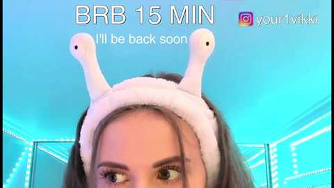 Video of a young woman with fair skin, brown eyes, and long brown hair, wearing a white, fluffy headband with two small, white, cartoonish ears. The background features a bright blue, lit room with white string lights. Text reads, \"BBR 15 MIN, I'll be back soon.\