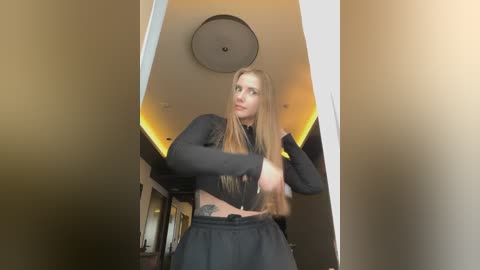 A video of a young woman with long, straight brown hair, wearing a black crop top and loose pants, standing in a dimly lit hallway with warm lighting. She has a tattoo on her side.