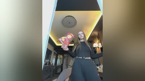 Video of a young woman with long brown hair, dressed in a black outfit, holding a red balloon in a modern, dimly lit room with a yellow ceiling light.