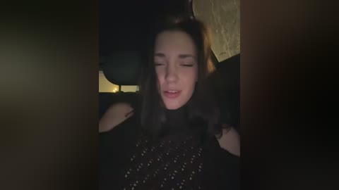 Video of a young woman with long, dark hair and light skin, wearing a black sequin dress, eyes closed, smiling, in a dimly lit room with a soft, golden light source.