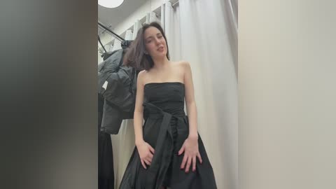 Video of a young Asian woman with shoulder-length dark hair, wearing a strapless black gown, standing in a fitting room with a white curtain and hanging clothes in the background.