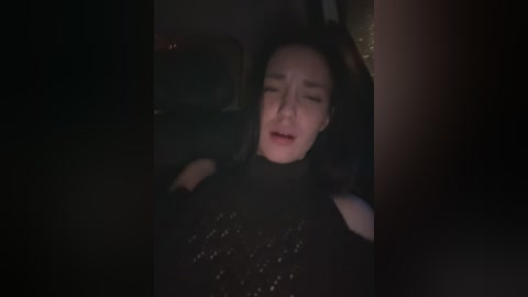 A dimly lit video of a woman with fair skin, long black hair, and closed eyes, wearing a black sequin dress, lying on a bed, with a blurred background.