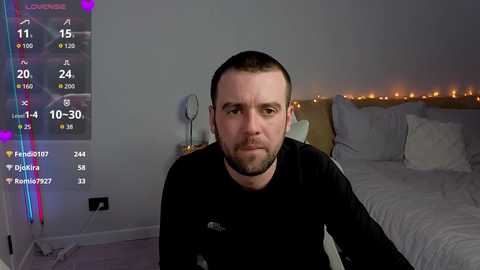 A video of a man with short brown hair and beard, wearing a black shirt, sitting on a bed with white sheets. The room has a dim, cozy atmosphere with fairy lights and a digital clock display.