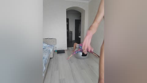 Video of a young person in a modern bedroom, wearing a blue and white patterned shirt, lying on a light-colored wooden floor. A hand, holding a red and blue object, is visible in the foreground.