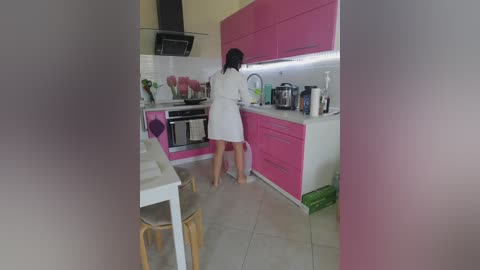 Video of a woman in a white dress standing at a bright pink kitchen counter, surrounded by modern appliances and pink cabinets, with a tiled floor and light walls.