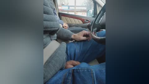 A video depicting a person wearing a gray sweater and blue jeans, seated in a car, buckling a seatbelt. The car interior shows beige upholstery, with a blurry background suggesting a street scene outside.