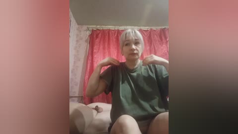 A video of an Asian woman with short gray hair, wearing a green t-shirt, sitting on a beige couch in a room with pink curtains and floral wallpaper.