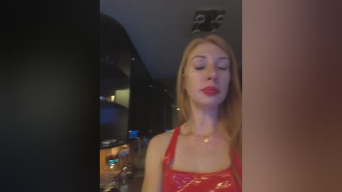 Video of a blonde woman in a red top, eyes closed, standing in a dimly lit room with a TV on the left and a black ceiling fan above.