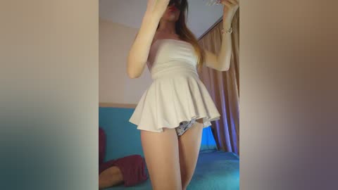 A video of a slim, fair-skinned woman with long brown hair, wearing a white, strapless, short dress that flares out, revealing patterned panties. She stands in a brightly lit room with blue and beige decor, holding a phone in front of her face.
