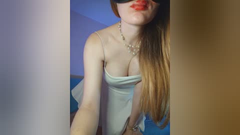 Video of a fair-skinned woman with long, straight, light-brown hair, wearing a white camisole and a black blindfold, red lipstick, and a delicate necklace, standing against a blue wall.