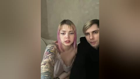 Video of a young woman with pink hair and a tattoo sleeve, sitting next to a man with short blonde hair, both looking serious in a dimly lit room.