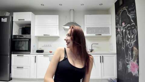 Video of a smiling woman with long, red hair in a black tank top, standing in a modern kitchen with white cabinets, a black fridge, and a chalkboard wall.
