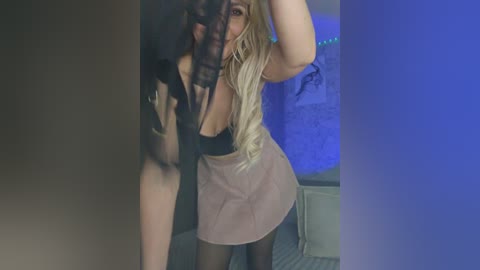 Video of a blonde woman with long hair, wearing a black bra and a short, light-colored skirt, holding a black mask over her face.