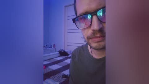 Video of a man with glasses, light skin, and trimmed beard, smiling slightly, in a dimly lit room with blue and purple lighting. Background shows a bed with striped bedding and scattered items.