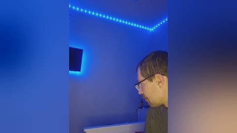 Video of a man with glasses, wearing a dark shirt, standing in a dimly lit room with blue LED strip lighting, looking at a TV screen mounted on a white wall.