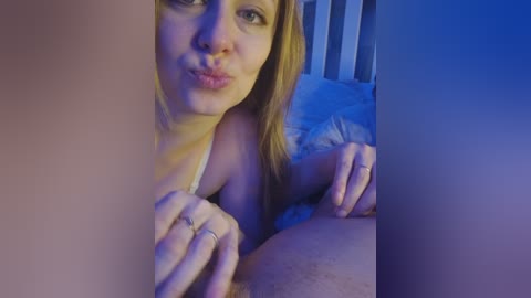 A video of a young, fair-skinned woman with light brown hair, wearing a white bra, making a kissy face, partially obscured by a dark object, in a dimly lit bedroom with blue lighting.