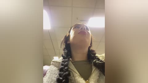 Video of a young woman with long black hair in braids, wearing a beige fleece jacket, standing in a dimly lit room with fluorescent lights on the ceiling.