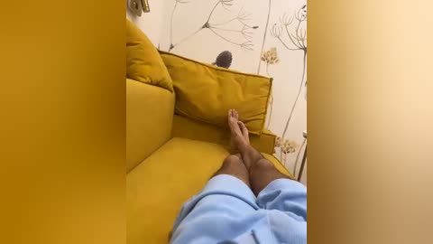 Video of a person with dark skin, wearing light blue shorts, lying on a mustard-yellow couch, feet up, against a white wall adorned with minimalist floral wallpaper.