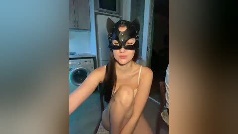 Video of a young woman with long hair, fair skin, and small breasts, wearing a black cat mask and beige bra, kneeling in a dimly lit laundry room with white cabinets and a washing machine.