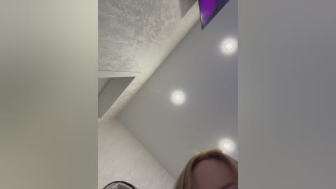 A video of a modern room with a white ceiling, recessed lighting, and a woman's head in the bottom right corner, partially cropped. The background features a textured, light gray wall and a flat-screen TV mounted on the wall.