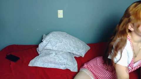 Video of a woman with long auburn hair in a white top and red checkered dress, lying on a red bed with a gray pillow and blanket, against a teal wall.