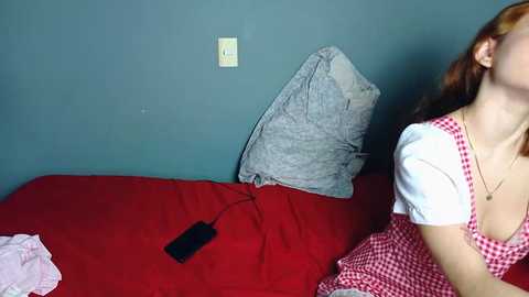 A video of a young woman with fair skin and red hair, wearing a red and white checkered dress, lying on a red bed with a gray pillow. A black device lies on the bed.