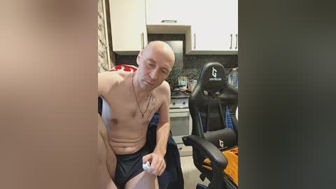 Video of a bald, muscular, fair-skinned man in black shorts, sitting shirtless in a kitchen, playing video games on a black gaming chair.