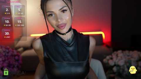 Video of a young woman with light skin, long black hair, and a black leather sleeveless top, sitting in a dimly-lit room with purple flowers and a red-lit background.