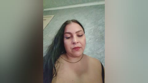 A video of a plus-sized, fair-skinned woman with long, dark hair, wearing a black bra and necklace, standing in a room with a textured, light blue wall and a wooden door visible in the background.