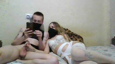 A video shows three naked people: a white man with a black mask, a white woman in white lingerie, and a tattooed man, all lying on a bed.
