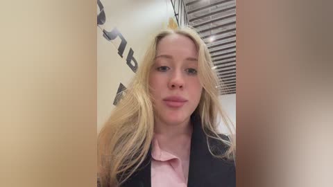 Video of a young blonde woman with long, wavy hair, wearing a pink blouse and black blazer, standing in an office with beige walls and partially visible blinds.