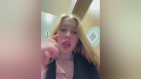 Video of a blonde woman with a shocked expression, wearing a pink blouse and black blazer, inside a narrow, beige-walled room with a white ceiling.