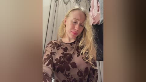 A video of a blonde woman with fair skin, wearing a sheer brown floral top, leaning against a beige wall with a partially open closet revealing pink and black clothing.