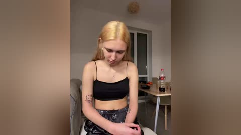 Video of a young, slender, blonde woman with fair skin, wearing a black spaghetti-strap crop top and high-waisted floral pants, seated in a chair in a modern, minimalist room with light walls and a wooden table.